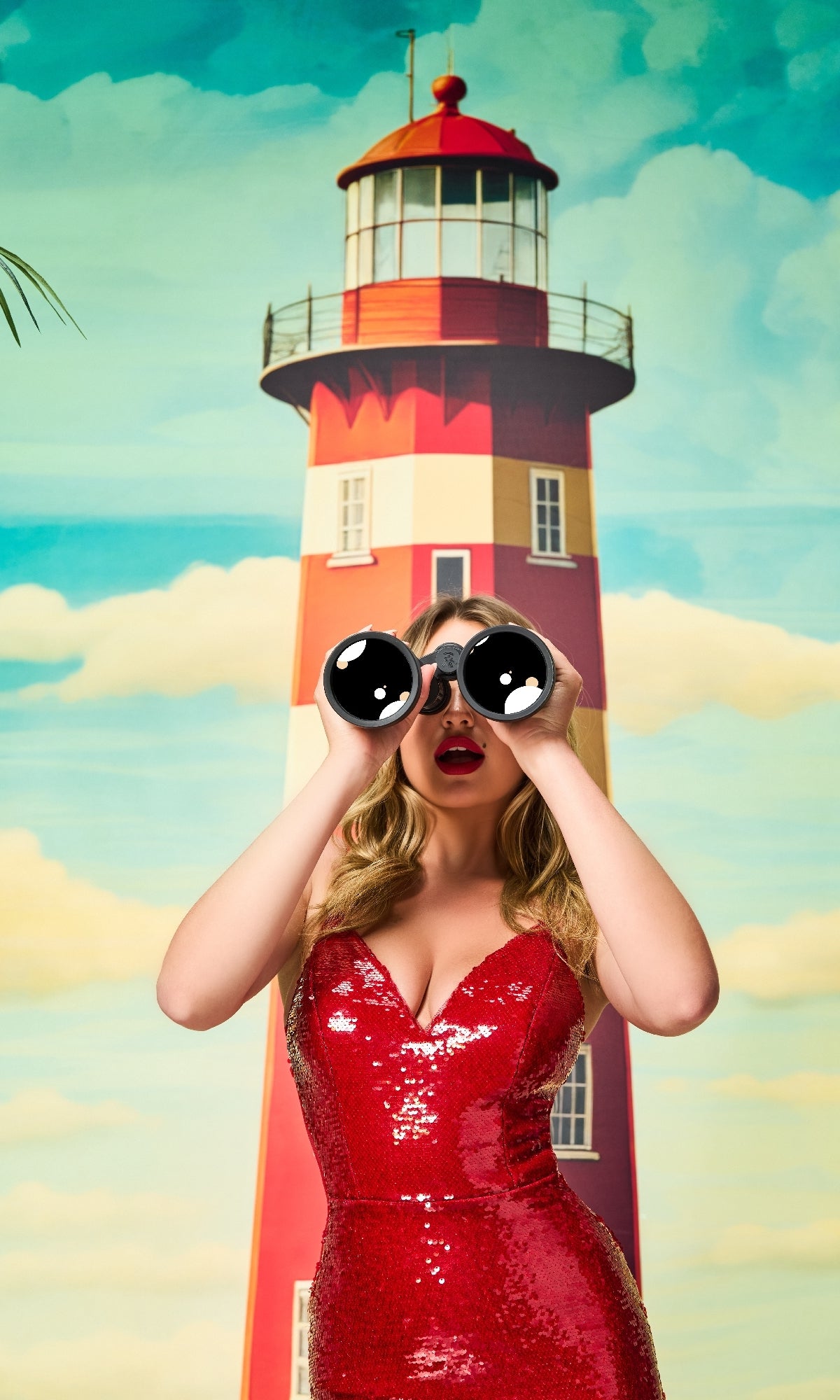 A woman in the Colette CL8615 long red sequin prom dress holds cartoon-eye binoculars near a red and white lighthouse, with a blue, cloudy sky in the background.